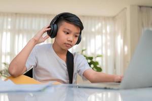 Asian boy playing guitar and watching online course on laptop while practicing for learning music or musical instrument online at home. Boy students study online with video call teachers play guitar. photo