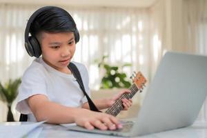 Asian boy playing guitar and watching online course on laptop while practicing for learning music or musical instrument online at home. Boy students study online with video call teachers play guitar. photo