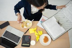 mujer de negocios ocupada y cansada comiendo espagueti para el almuerzo en la oficina y trabajando para entregar estados financieros a un jefe. con exceso de trabajo y poco saludable para comidas preparadas, concepto de agotamiento. foto