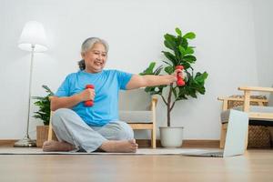 Asian senior woman lifting dumbbell for exercise and workout at home. Active mature woman doing stretching exercise in living room. Exercise Active and healthy for older, elder, and senior concept. photo