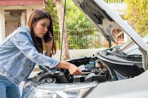 mujer asiática enojada y usando un teléfono móvil pidiendo ayuda después de una avería en el coche en la calle. concepto de problema del motor del vehículo o accidente y ayuda de emergencia de un mecánico profesional foto