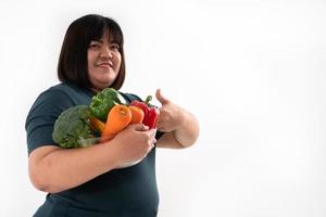 feliz mujer asiática con sobrepeso levanta el pulgar, sosteniendo una canasta de verduras y sonriendo sobre un fondo blanco aislado, el concepto de buena salud proviene de comer una dieta nutritiva. foto