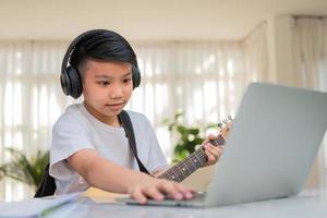 Asian boy playing guitar and watching online course on laptop while practicing for learning music or musical instrument online at home. Boy students study online with video call teachers play guitar. photo