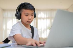 Asian boy playing guitar and watching online course on laptop while practicing for learning music or musical instrument online at home. Boy students study online with video call teachers play guitar. photo