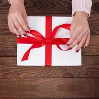Woman's hands wrapping christmas, holiday handmade present in paper box with red ribbon. Give present box, decoration of gift on vintage wooden table, top view, copy space mock up template for design. photo