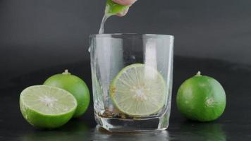 mano exprimiendo lima en un vaso con hielo y rodajas de limón con limas alrededor de una mesa de fondo negro. bebida alcoholica. video
