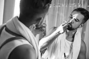 Man looking in the mirror, smoking a cigarette and shaving his beard with a straight razor photo