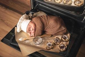 Newborn baby wearing chef's hat is lying on the oven tray with a muffins photo