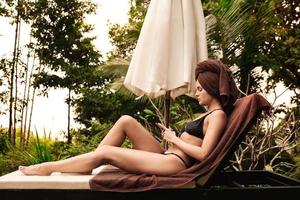 Woman relaxing and using smartphone on wooden sunlounger after bathing in swimming pool photo