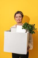 Upset woman holding box with personal items after job resignation against yellow background photo