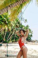 Gorgeous female model wearing red bikini near swing on the sandy beach with a lot of coconut palms photo