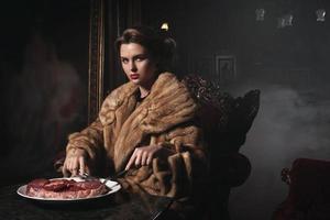 Provocative image of woman wearing wearing fur coat and eating piece of raw beef. Animal rights concept. photo