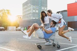tres amigos diversos divirtiéndose y montando carrito de compras en el estacionamiento cerca del supermercado