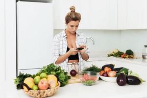 mujer con auriculares inalámbricos está usando un teléfono inteligente durante la cocina en una cocina blanca moderna foto