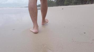 laag hoek visie langzaam beweging van man's poten en blootsvoets wandelen langs zee water golven Aan zanderig wit strand.wandelingen Bij kust surfen. spatten van water en schuim in langzaam beweging. video