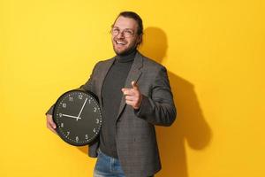 Cheerful man wearing glasses holding big clock on yellow background photo