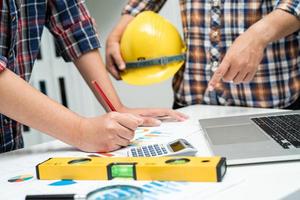 Asian engineer and partner team learn and talk with house plan project blueprint, graph and yellow helmet and engineering construction tools on table work. photo