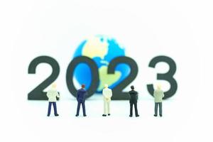 2023 Global Business New Year Concept. Closeup of group of businessman miniature figures standing and looking to number  and mini world ball on white background. photo