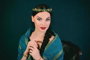 Portrait of young beautiful brunette in Indian style with wreath and braid in a blue scarf and gold bracelets on dark background photo