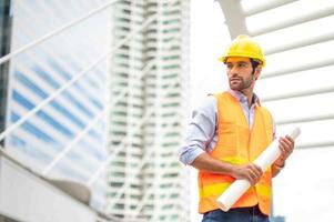 Young caucasian man holding a big paper, guy wearing light blue shirt and jeans with orange vest and yellow helmet for security in construction area. photo