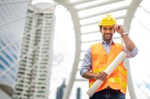 Young caucasian man holding a big paper, guy wearing light blue shirt and jeans with orange vest and yellow helmet for security in construction area. photo