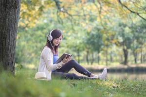 Young Asian college student is listening to audio book while sitting under the tree by the lake at the university campus natural park for education and relaxation in nature concept photo