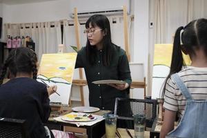 A female Asian teacher teaches and demonstrates to the children on acrylic color picture painting on canvas in art classroom, creatively learning with skill at the elementary school studio education. photo