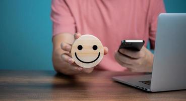 Man using Mobile Phone and holding wooden disc with smiling face icons, customer satisfaction service, feedback, rating and positive customer review, experience, world mental health day. photo