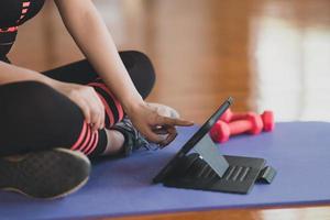 Crop picture. Hand's sportswoman touching on tablet sitting on yoga mat and doing exercise with dumbbell workout online and looking video streaming on tablet at fitness gym. Online fitness concept. photo