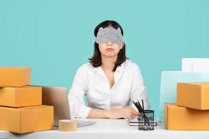 Tired and sleepy young Asian businesswoman in a casual shirt and using sleep mask blindfold sitting at a white desk with laptop and brown cardboard isolated on pastel green background. photo