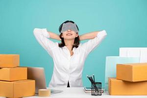 Relaxing Asian businesswoman sitting at her desk with laptop and brown cardboard isolated on pastel green background. She put her free hand on the nape of her neck. photo