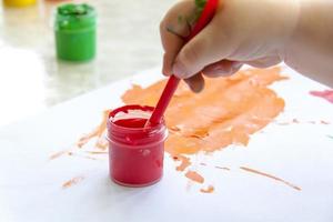 A child's hand holds and dips the brush in the paint. Smears of bright paint on white paper. photo