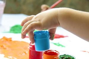 A child's hand holds a jar of paint about to draw with it. Smears of bright paint on white paper. photo