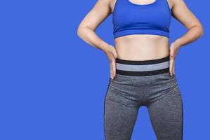 Fitness girl in a top and leggings points to her athletic figure. The Concept Of Weight Loss. Model on a blue background. A slender Young Lady Holds her hands at her waist. photo