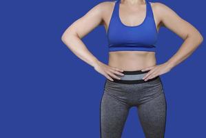 Fitness girl in a top and leggings points to her athletic figure. The Concept Of Weight Loss. Model on a blue background. A slender Young Lady Holds her hands at her waist. photo
