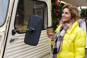 Happy woman with a cup of hot coffee beside the retro van at Christmas market photo