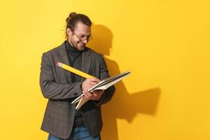 Cheerful man wearing glasses holding big pencil and notebook on yellow background photo