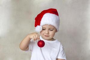 Portrait of a cute little boy in a white t-shirt and Santa Claus hat. A boy admires a red Christmas ball. Children's emotions. Christmas and new year. photo