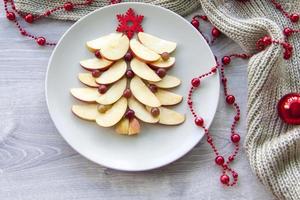 Christmas tree made of sliced apples and berries with a large red wooden snowflake instead of a star. Plate with apples on a light background. photo
