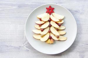 Christmas tree made of sliced apples and berries with a large red wooden snowflake instead of a star. Plate with apples on a light wooden background. photo