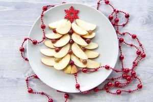 Christmas tree made of sliced apples and berries with a large red wooden snowflake instead of a star. Plate with apples on a light wooden background. photo