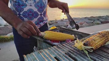 Le vendeur de nourriture de rue au bord de l'océan prépare des épis de maïs sur des images de barbecue. video