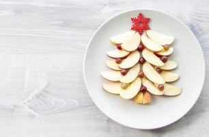 Christmas tree made of sliced apples and berries with a large red wooden snowflake instead of a star. Plate with apples on a light wooden background photo