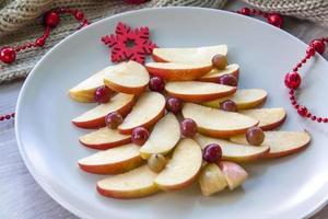 Christmas tree made of sliced apples and berries with a large red wooden snowflake instead of a star. Plate with apples on a light background. photo