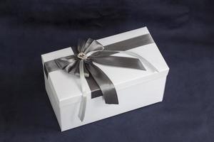 la caja de regalo es blanca con un hermoso lazo gris. regalo sobre un fondo oscuro. vacaciones y sorpresas. lazos de satén con pedrería. foto