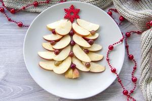 Christmas tree made of sliced apples and berries with a large red wooden snowflake instead of a star. Plate with apples on a light background. photo