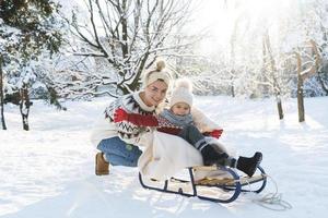 Young mother and her cute little son with retro sled in a snowy park during sunny day photo