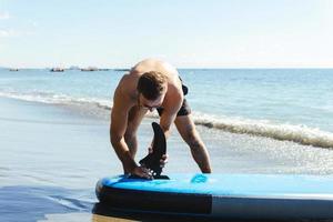 Young male surfer setting up standup paddleboard on a beach. photo