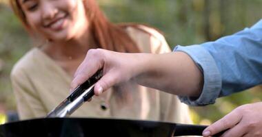 Selective focus hand of young asian woman cooking and her friend enjoy to make the meal in pan, They are talk and laugh with fun together while camping in nature park video