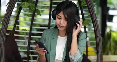 bella giovane asiatico donna seduta e godere per ascoltando musica a partire dal senza fili cuffie con smartphone a bar. le persone, rilassante e stile di vita concetto. video